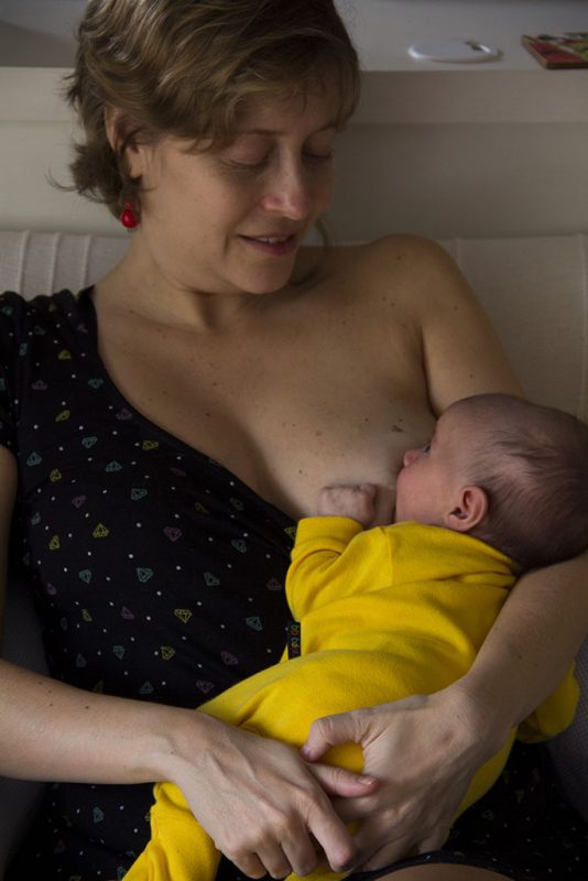 Foto alternativa de mulher amamentando o filho. Ela aparece com a criança no colo e olha para ela.