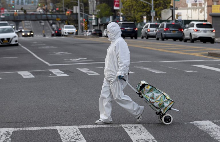 Uma mulher vestindo traje e óculos de proteção puxa seu carrinho de compras nas ruas de Queens, em Nova York. Foto Johannes Eisele/AFP