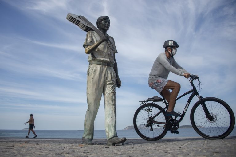 Homem usando máscara pedala ao lado da estátua do maestro Tom Jobim, no Arpoador, Rio de Janeiro. Foto Mauro Pimentel/AFP