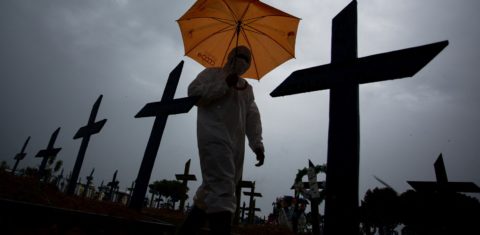 Trabalhador com traje de proteção e guarda-chuva passa por entre os túmulos das vítimas da covid-19 no cemitério de Nossa Senhora Aparecida, em Manaus. Foto Michael Dantas/AFP. Fevereiro/2021