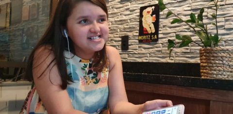 Fotografia da Isabela segurando celular com a mão esquerda, com fones de ouvido conectados para audiodescrição. No celular, a tela mostra a interface do aplicativo Vem Cá. Ela tem expressão sorridente