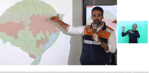 Captura de tela colorida do governador do Rio Grande do Sul Eduardo Leite. Leite está de pé ao lado de um mapa do RS com partes em vermelho nas regiões central e metropolitana de Porto Alegre. Ele segura um microfone e aponta com a mão direita para o mapa. Ele usa um colete da defesa civil e uma camiseta branca por baixo. Leite é um homem branco com barba e cabelos pretos. Ao seu lado, em uma tela pequena aparece uma intérprete. Ela é uma mulher negra com uma camiseta preta e está sinalizando em Libras, em um fundo verde-azulado.