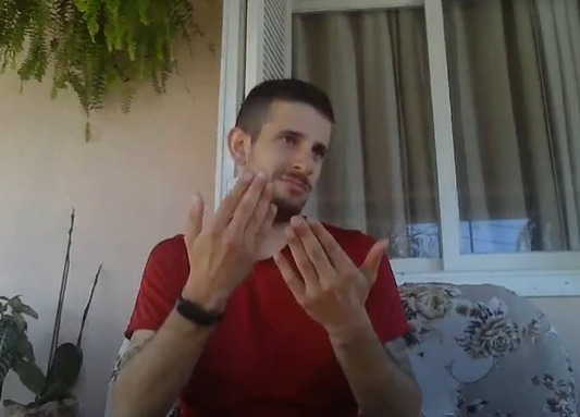 Captura de tela colorida de Andrei sinalizando. Ele é um homem branco com cabelo curto e barba preta. Ele usa uma camiseta vermelha e sinaliza com as mãos na frente do corpo, enquanto olha para o lado. Ao fundo, janela e parede de sua casa.