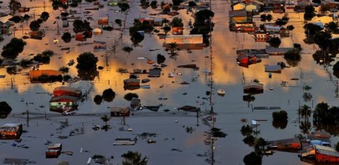 Foto colorida da região Metropolitana de Porto Alegre submersa após enchente. A água toma a maior parte da imagem, com apenas alguns pontos em que aparecem árvores e casas não inundadas.