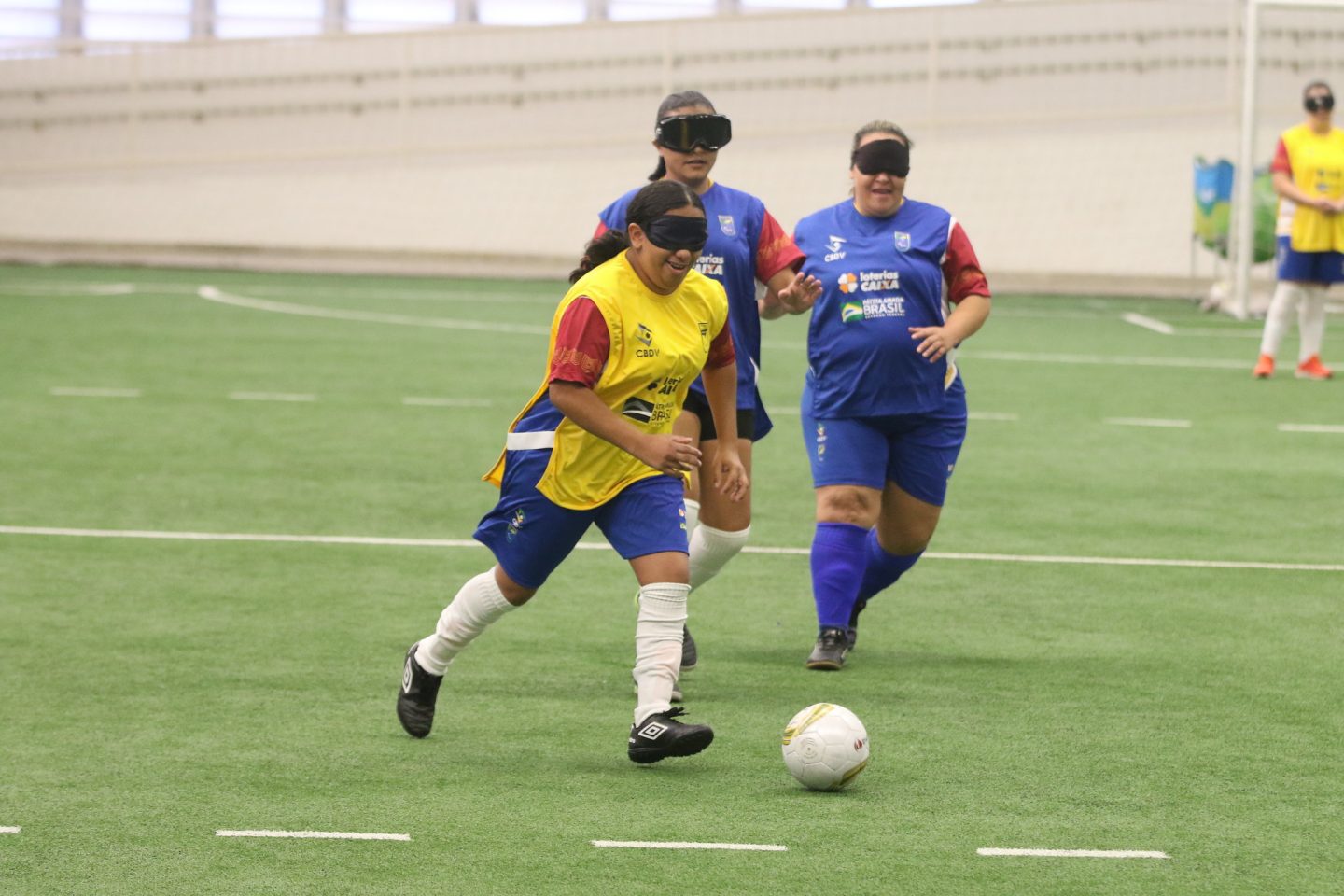 Foto colorida de atletas disputando jogo de futebol de cegas em São Paulo. No centro da imagem, uma mulher cega de camiseta amarela corre com a bola, enquanto sorri. Ela usa bermuda azul e meias brancas, além de proteção nos olhos. Ao fundo, aparecem duas mulheres vestidas de camiseta, bermuda e meias azuis, elas correm em direção à bola.