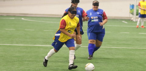 Foto colorida de atletas disputando jogo de futebol de cegas em São Paulo. No centro da imagem, uma mulher cega de camiseta amarela corre com a bola, enquanto sorri. Ela usa bermuda azul e meias brancas, além de proteção nos olhos. Ao fundo, aparecem duas mulheres vestidas de camiseta, bermuda e meias azuis, elas correm em direção à bola.