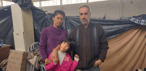 Foto colorida de Adelar, Valeria e Pamela em abrigo de Lajeado. Eles estão abraçados e usam roupa de frio, ao fundo barraca de abrigo.