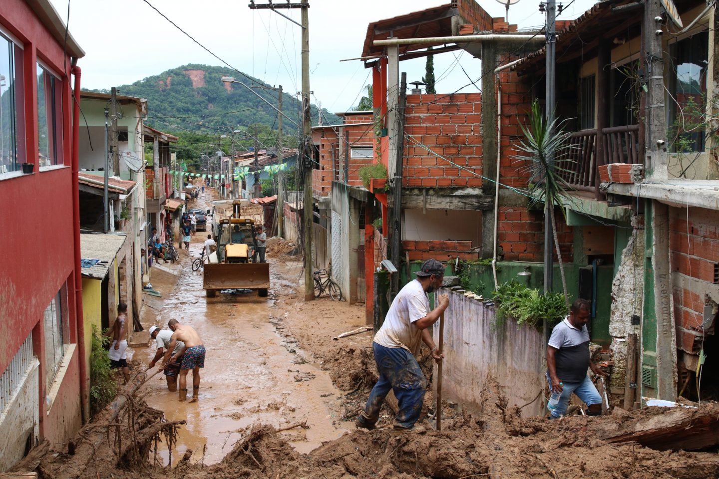 Foto colorida a Vila Sahy após desastre. Na imagem, aparecem casas destruídas e a rua tomada por lama, enquanto pessoas trabalham para limpar o local