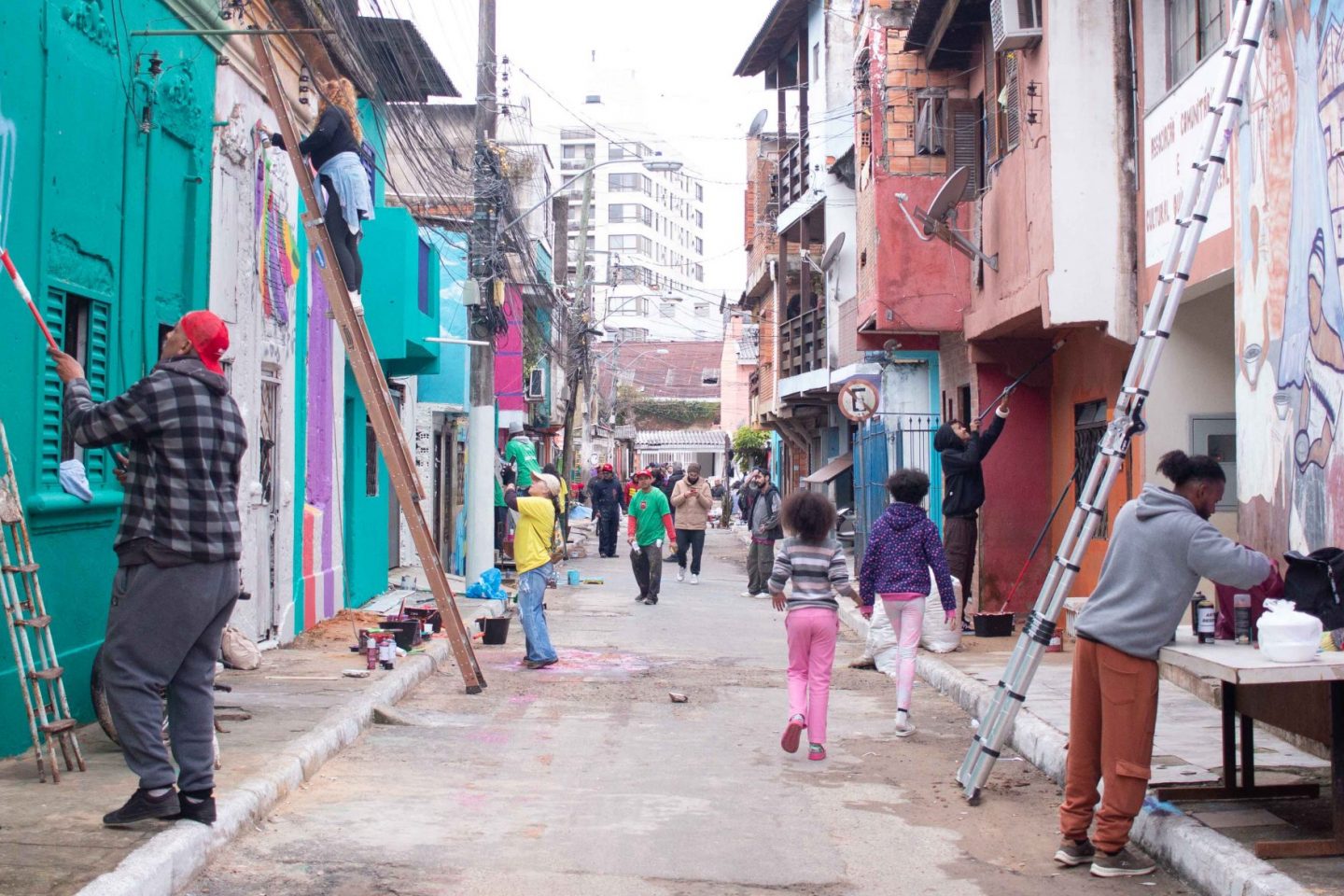 Foto colorida de mutirão do Paredes com Proposito. Na imagem, diversas pessoas aparecem pintando residências em rua do Quilombo do Areal, em Porto Alegre. Artistas pintam paredes, enquanto moradores passeiam pelo local