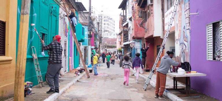Foto colorida de mutirão do Paredes com Proposito. Na imagem, diversas pessoas aparecem pintando residências em rua do Quilombo do Areal, em Porto Alegre. Artistas pintam paredes, enquanto moradores passeiam pelo local