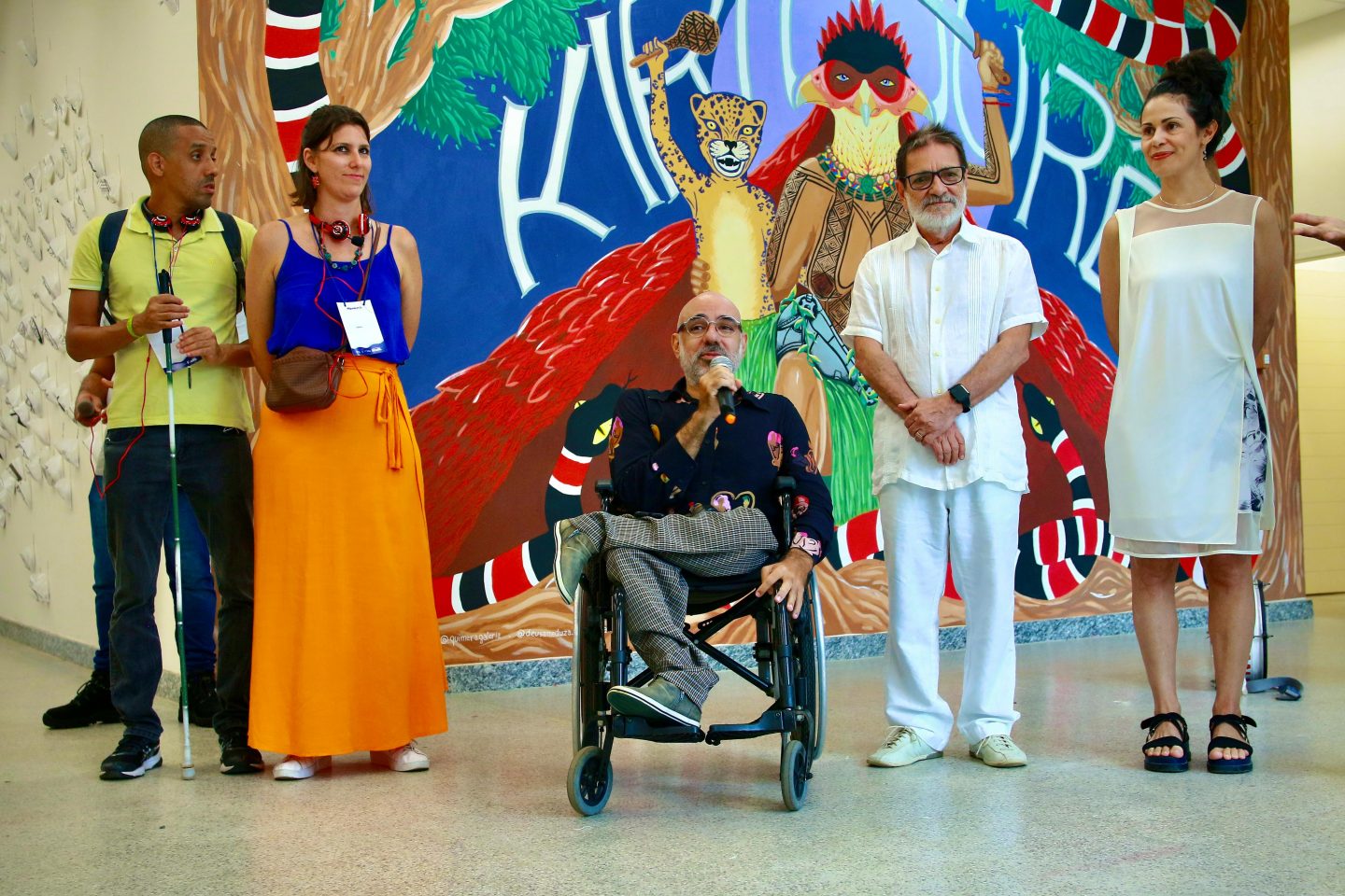 Foto colorida de Edu O. junto ao Reitor da UFBA - prof Paulo Miguez, da profa Rita Aquino representante da PROEXT-BA (Pro-Reitoria de Extensão) e da equipe de audiodescrição Jurema Iris e Deivide Monteiro, durante lançamento do mapeamento acessa mais. Edu é um homem branco, usuário de cadeira de rodas. Ele está com o microfone na mão. Ao fundo, parede com arte colorida