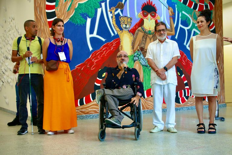 Foto colorida de Edu O. junto ao Reitor da UFBA - prof Paulo Miguez, da profa Rita Aquino representante da PROEXT-BA (Pro-Reitoria de Extensão) e da equipe de audiodescrição Jurema Iris e Deivide Monteiro, durante lançamento do mapeamento acessa mais. Edu é um homem branco, usuário de cadeira de rodas. Ele está com o microfone na mão. Ao fundo, parede com arte colorida