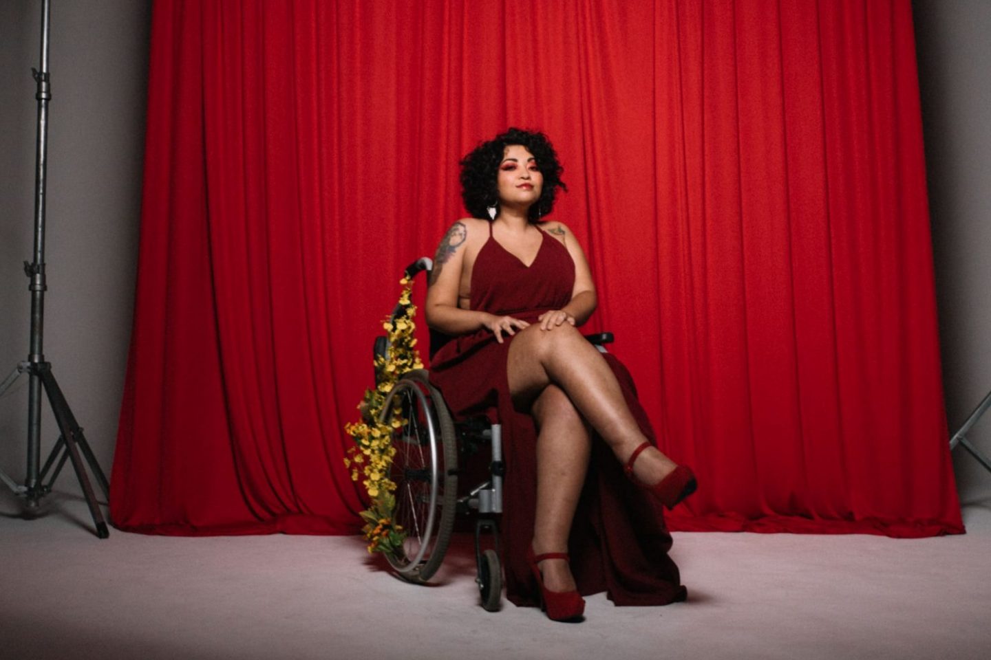 Foto colorida da artista def Amanda Lyra. Ela é uma mulher branca com cabelos cacheados pretos. Ela usa um vestido vermelho e está em uma cadeira de rodas enfeitadas com flores amarelas. Ao fundo, cortina vermelha.