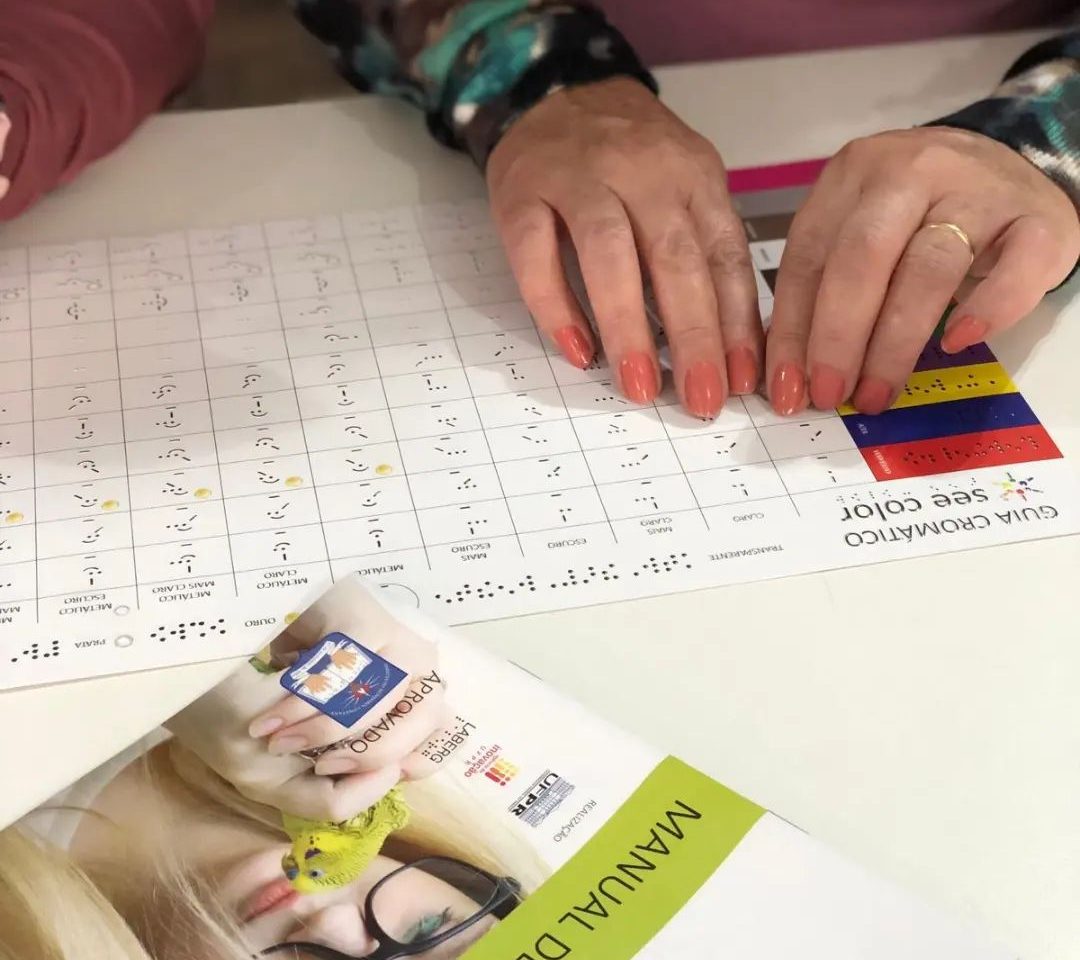 Foto colorida das mãos de uma pessoa sob uma folha com códigos de cores em braille e no See Color