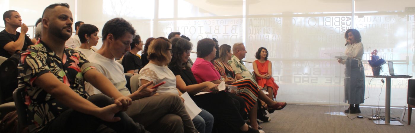 foto colorida do lançamento da biblioteca virtual Mais Diferenças. Á esquerda, diversas pessoas assistem a apresentação da plataforma. À direita da imagem, Lara Souto, uma mulher negra com cabelos cacheados e usando uma blusa branca e saia cinza, fala em um microfone. Ela está diante de um púlpito transparente. Ao fundo, parede branca.