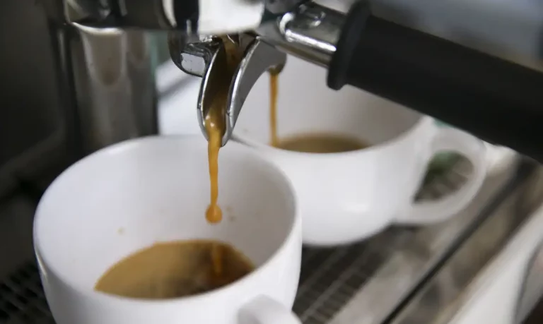 Foto colorida de xícara de café sendo enchida. Na imagem, parte da bebida aparece nas xícaras brancas e parte caindo de torneira