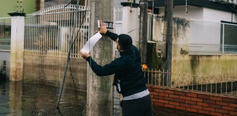 Foto colorida de pessoa colocando adesivo do Água Até Aqui em poste, em meio a inundação