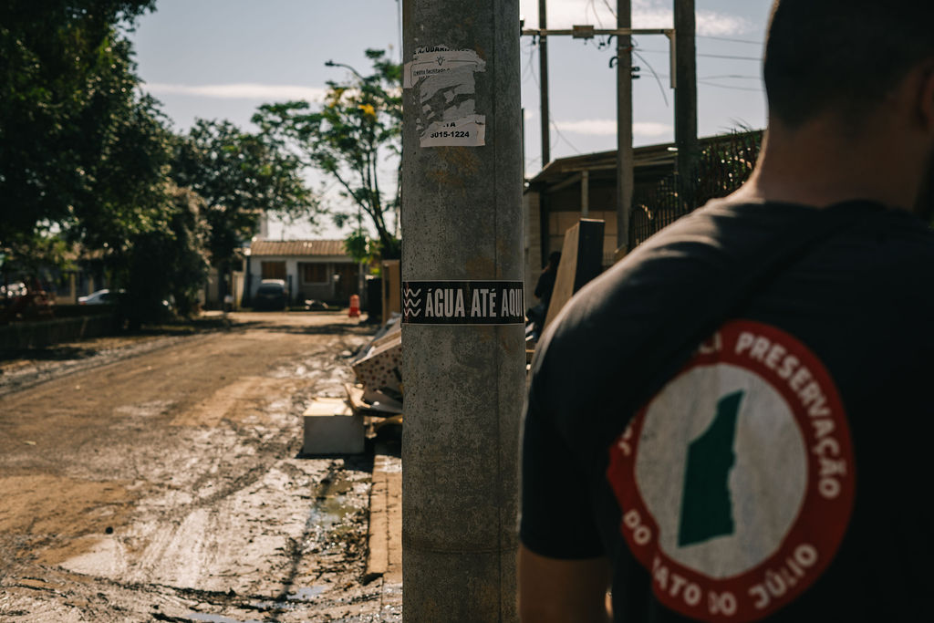 Foto colorida de adesivo do Água até aqui colocado em poste. Ao fundo, casas cobertas por lama. E no primeiro plano uma pessoa de costas