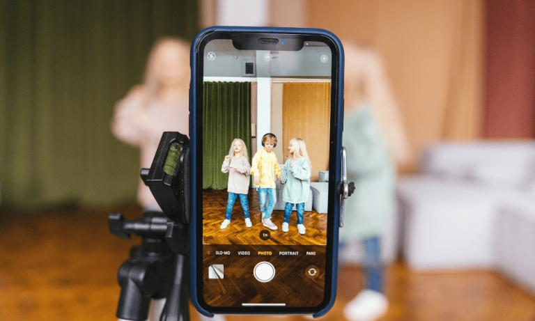 Foto colorida de uma tela de celular posicionada para filmar crianças em pé. Prática de sharenting pode expor menores de idade