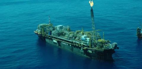 Foto colorida de plataforma de petróleo operando no mar. Em uma das torres, é possível ver uma chama da queima de combustíveis