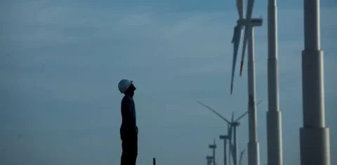 Foto colorida do perfil de um homem de pé diante de turbina de energia eólica. Ao fundo, céu azul