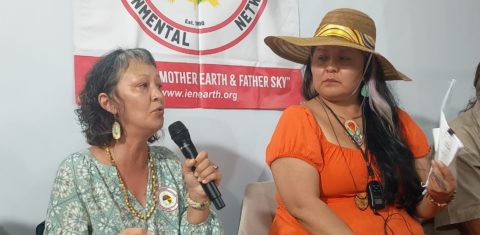 Foto colorida de Aakaluk, nome nativo de Adrienne La-Ree Blatchford. Ela é uma mulher indígena de cabelos curtos pretos e meio grisalhos. Ela está com um vestido verde e segura um microfone