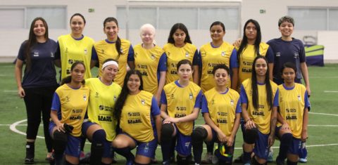 Foto colorida de um grupo de mulheres em um campo de futebol coberto. Elas estão posando organizadas em duas fileiras: a fileira de trás está em pé e a da frente está ajoelhada. A maioria das mulheres veste uniforme esportivo com colete amarelo, com o logo da Loterias Caixa, Governo Federal e Brasil estampados. Algumas usam faixa na cabeça e joelheiras. Nenhuma das atletas está com o óculos de proteção no momento da foto. No fundo, há um gol e uma parede branca com janelas e a inscrição "Secretaria dos Direitos da Pessoa com Deficiência - São Paulo". O ambiente é iluminado e o campo tem gramado sintético. Crédito: Renan Cacioli/ CBDV.