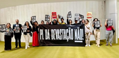 Foto colorida mostra pessoas em manifestação contra o PL da Devastação que implodiu o licenciamento ambiental no Brasil. Elas seguram uma faixa preta com letras brancas que diz: PL da Devastação não!