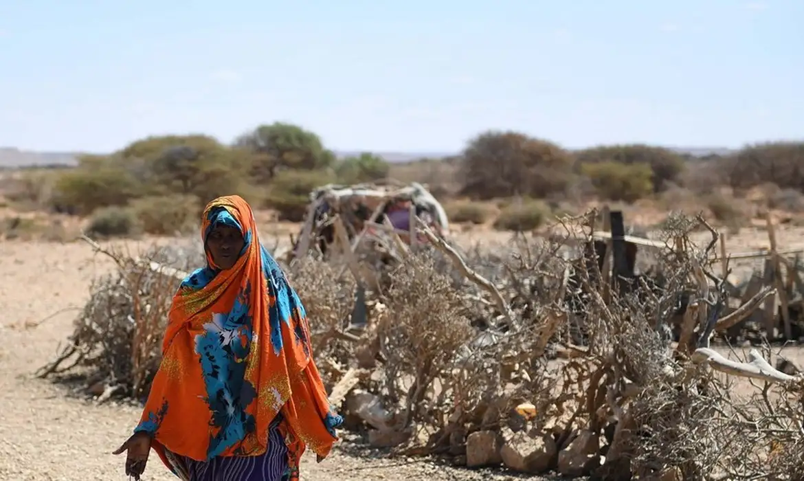 Foto colorida de uma mulher preta coberta com vestes azuis e laranjas andando em local afetado por seca na África Oriental