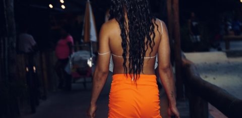 Foto colorida de Nina 100 Babado de costas. Ela é uma mulher trans negra, tem cabelo preto, longo ondulado. Ela usa um vestido laranja