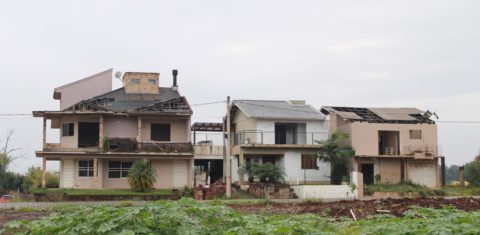 Foto colorida que mostra três casas afetadas pelas enchentes no Bairro Navegantes, em Arroio do Meio. As casas apresentam diversas avarias e buracos nos telhados, além de paredes quebradas