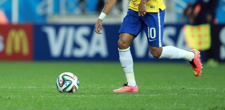 Foto colorida que mostra bola de futebol e jogador da seleção brasileira em partida da Copa do Mundo de 2014. Na imagem, o jogador aparece apenas da cintura para baixo e se desloca em direção à bola