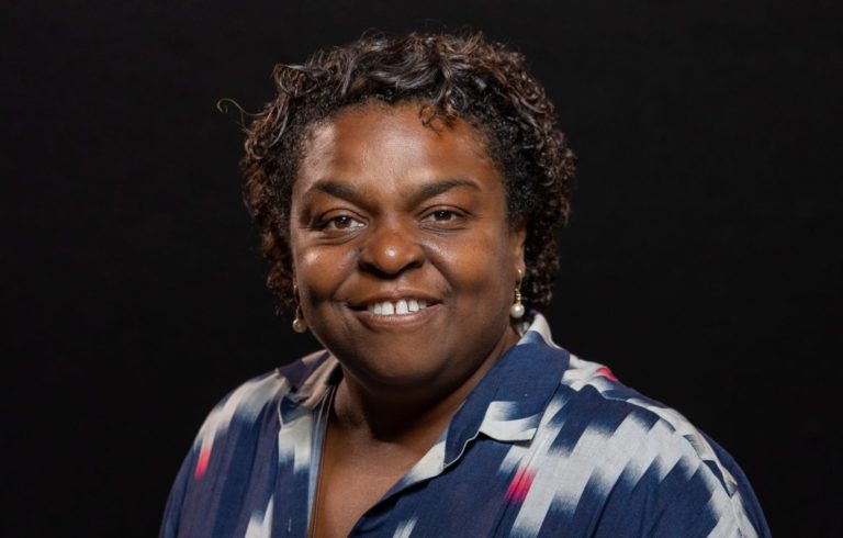 Foto colorida de Andrea Pitta. Ela é uma mulher negra, com cabelo curto, preto. Ela está usando uma blusa azul com detalhes cinza e sorri
