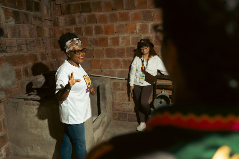 Foto colorida que mostra a mestra Zeza do Coco, no canto esquerdo. Ela é uma mulher negra, com cabelo afro e usa óculos e uma camiseta branca, enquanto gesticula com a mão direita