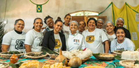 Foto colorida com grupo de mulheres da Associação de Mulheres Guerreiras Quilombolas do Castainho. Elas são quilombolas, negras, usam uma camiseta branca da associação e estão atrás de uma mesa com várias comidas feitas a partir de mandioca