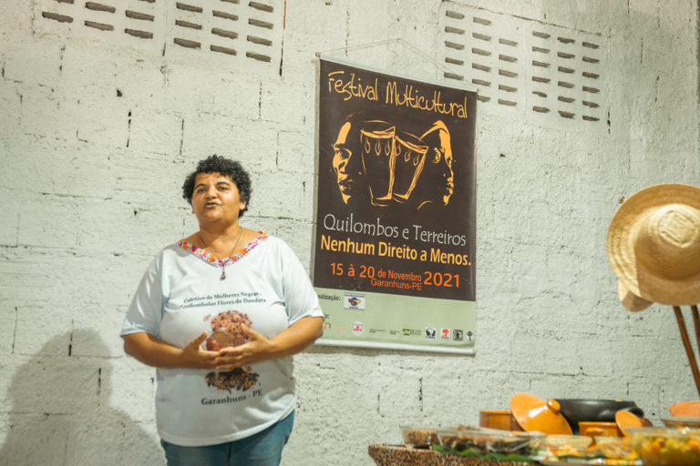 Foto colorida de Aparecida Nascimento Oliveira. Ela é uma mulher negra, quilombola, e está de pé, usando uma camiseta branca. Ela tem cabelo curto, preto.