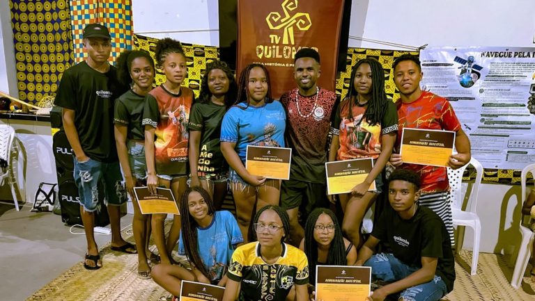 Foto colorida de vários jovens quilombolas ao lado de banner da Tv Quilombo. Eles seguram um certidicado e sorriem