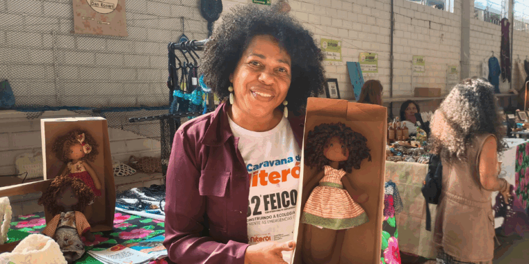 Foto colorida de Cristina Santos. Ela é uma mulher negra, com cabelo cacheado preto e segura uma boneca de pano negra.