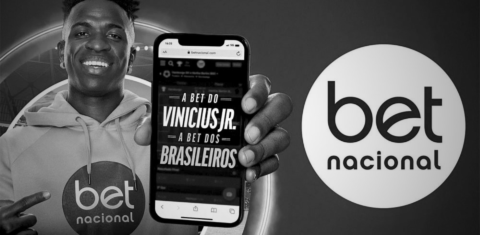 Imagem em preto e branco com o jogador Vinícius Jr fazendo propaganda de bet. Ele aparece com a camiseta da casa de aposta e segurando um celular
