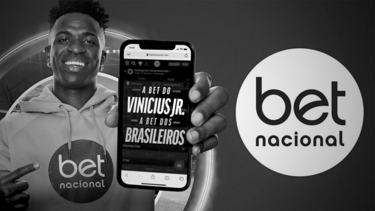 Imagem em preto e branco com o jogador Vinícius Jr fazendo propaganda de bet. Ele aparece com a camiseta da casa de aposta e segurando um celular