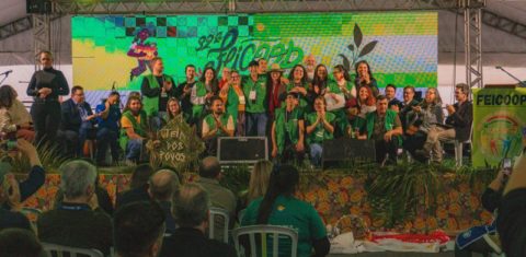 Foto colorida do palco da 32° Feicoop, durante cerimônia de abertura do evento. Na imagem, aparecem diversas pessoas da organização reunidas no palco. Evento discute impacto da crise climática na economia solidária