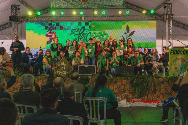 Foto colorida do palco da 32° Feicoop, durante cerimônia de abertura do evento. Na imagem, aparecem diversas pessoas da organização reunidas no palco. Evento discute impacto da crise climática na economia solidária