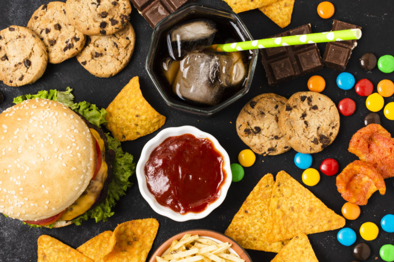 Foto colorida com vários alimentos ultraprocessados em cima de uma mesa. Na imagem, aparece um hamburguer, biscoitos, refrigerante e salgadinhos