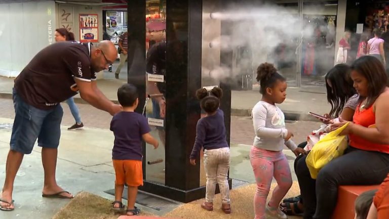Foto colorida de pessoas em torno de refúgio climático em Belo Horizonte. Na imagem, nuvens de nebulização aparecem em volta de crianças e adultos