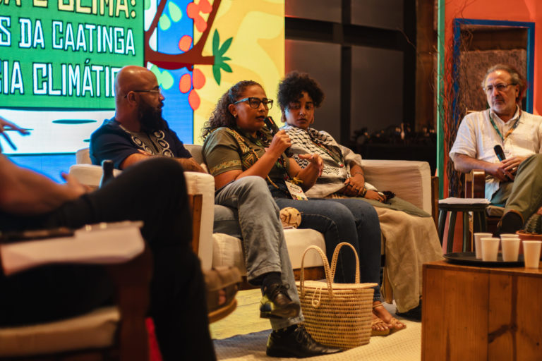 Foto colorida de painel de abertura da Caatinga Climate Week. Na imagem, Romana Kambiwá aparece com o microfone. Ela é uma mulher indígena, com cabelo preto e usa óculos. Ela está sentada ao lado de Antônio Crioulo e Marcele Oliveira