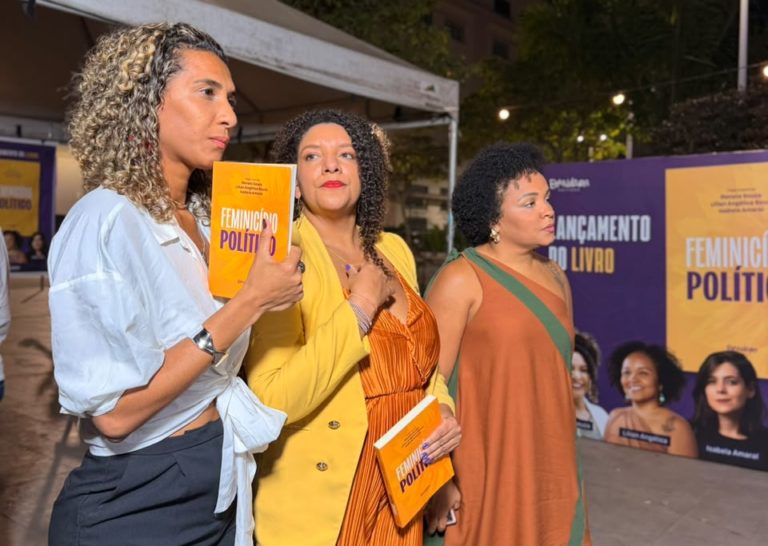 Foto colorida de Anielle Franco, ao lado de Renata Souza e de Lilian Angélica. Elas são três mulheres negras e estão segurando o livro "Feminicídio político", em evento de lançamento no Rio de Janeiro