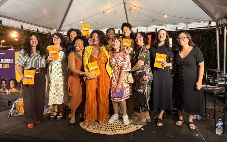 Foto colorida de Renata Souza ao lado de outras autoras do livro "Feminicídio político". Elas sorriem e estão de pé, vários seguram o livro nas mãos