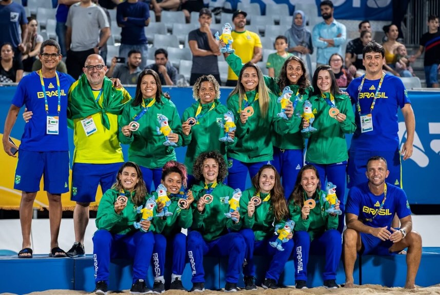O técnico Fabrício Santos com as jogadoras da seleção feminina e suas medalhas de bronze do Beach Games 2021: buscando abrir caminhos no beach soccer para as mulheres (Foto: Foto: Manuel Queimadelos / BSWW - 15/08/2021)